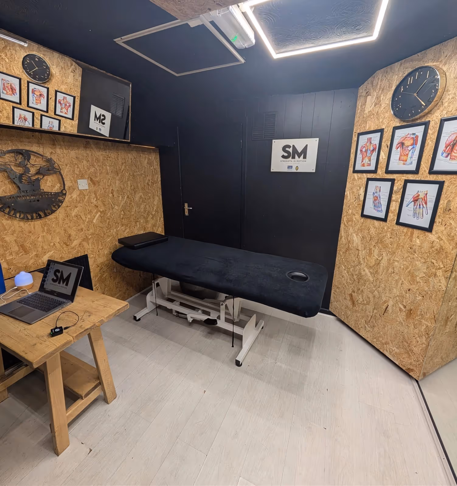 Therapy or massage room with a black treatment table, wooden desk with a laptop, anatomical muscle charts on the walls, and two clocks.