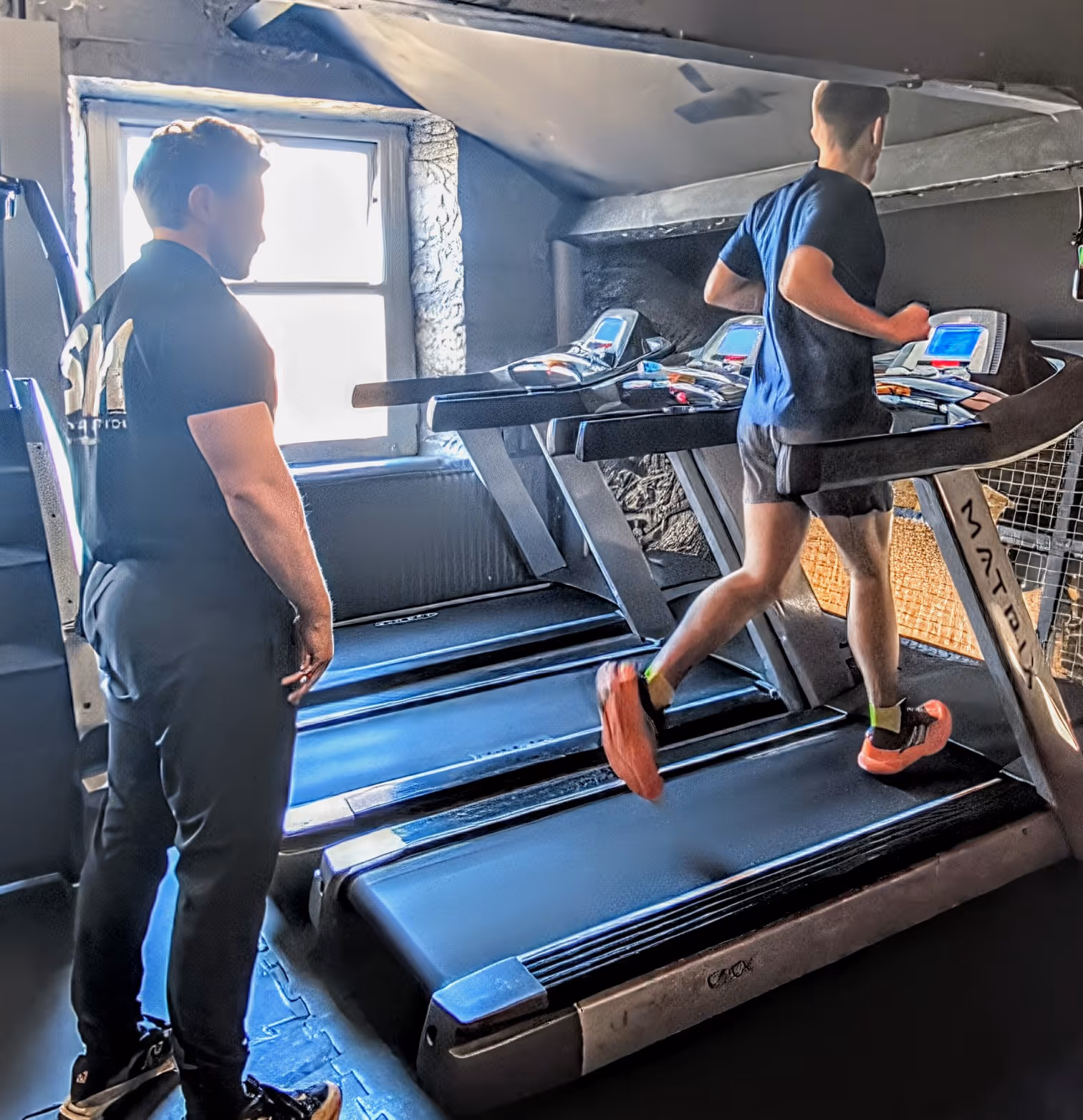 A man runs on a treadmill in a gym while another man, wearing a black shirt and pants, stands nearby watching.