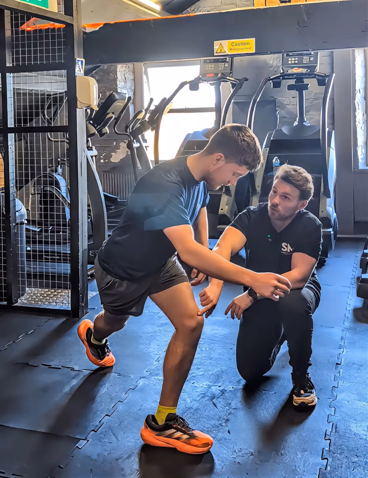 Gym trainer instructing a young man on lunge exercise technique indoors.