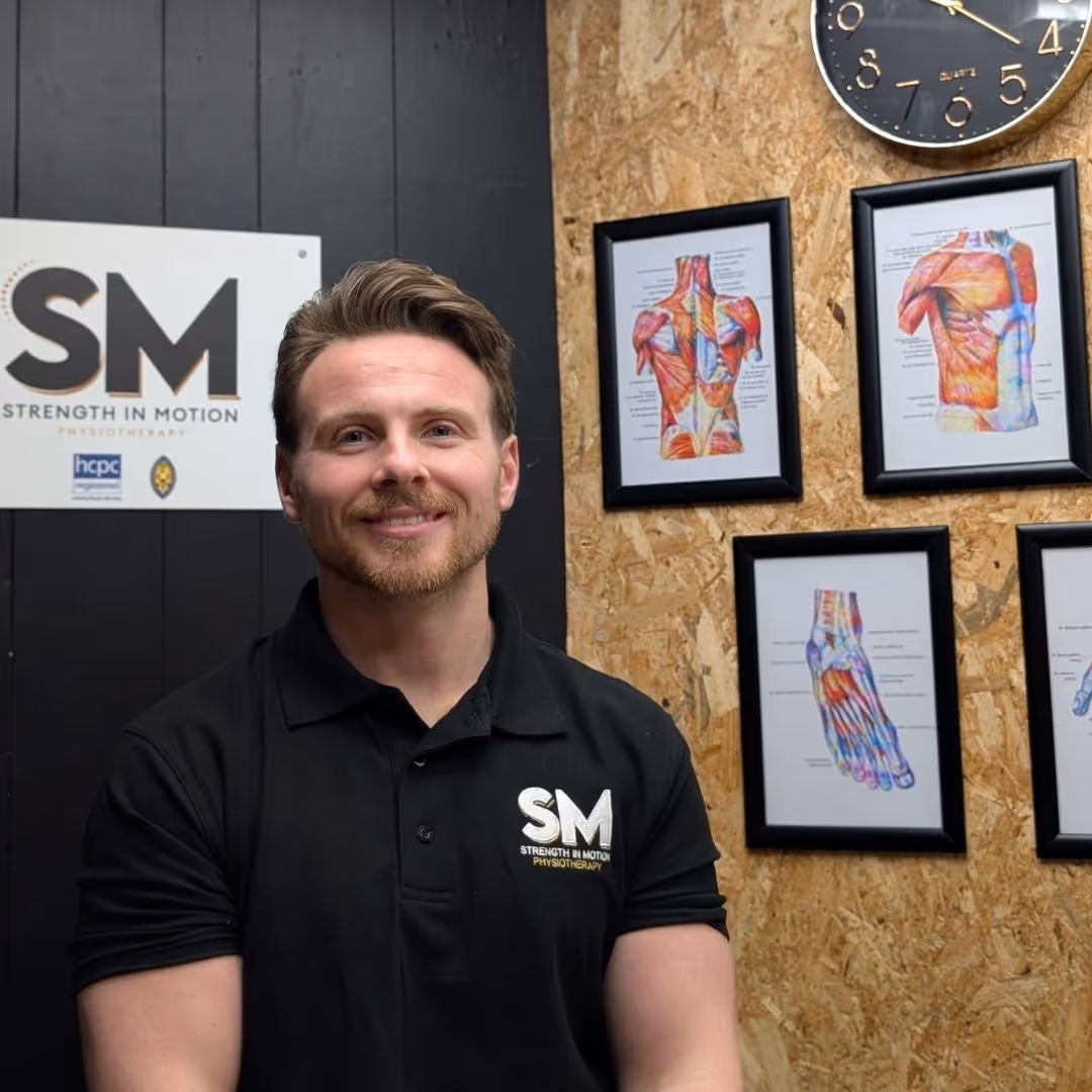 Smiling man in a black polo shirt with 'SM Strength in Motion Physiotherapy' logo standing in front of a wall with framed anatomical muscle charts and a clock.