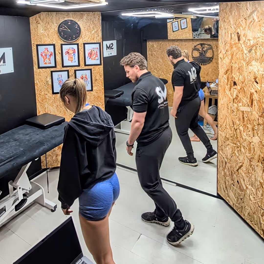 A male instructor in black guides a female athlete in blue shorts and a black hoodie performing a stretching exercise in a gym room with anatomical posters and a large mirror.