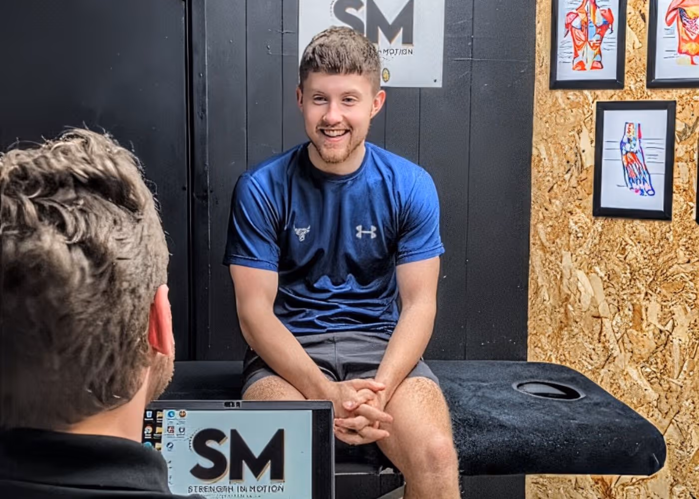 Young man in a blue Under Armour shirt sitting on a black examination table smiling during a consultation with a person using a laptop displaying 'Strength In Motion'.