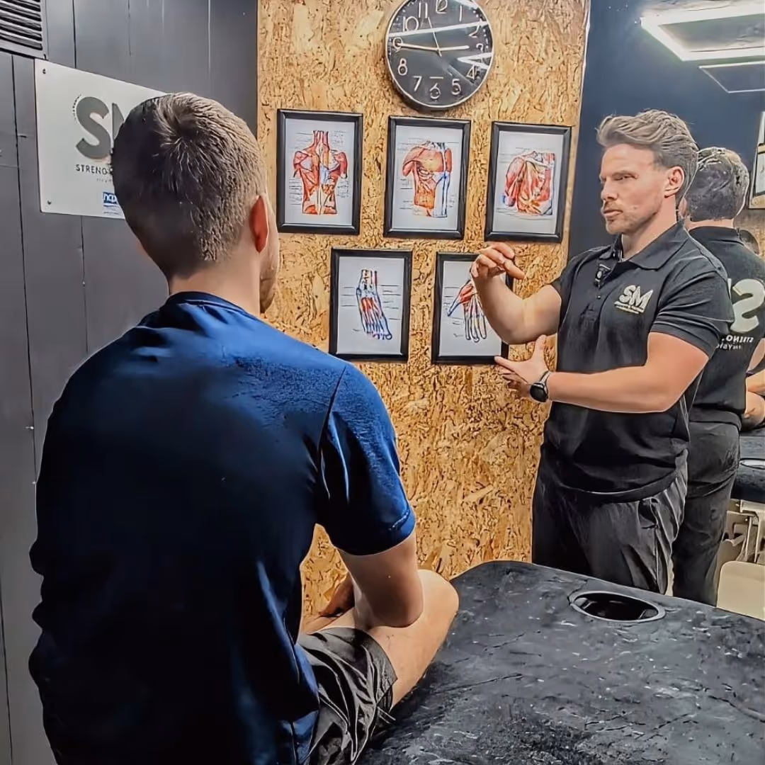 Physiotherapist explaining hand muscle anatomy to a seated patient using framed diagrams on the wall.